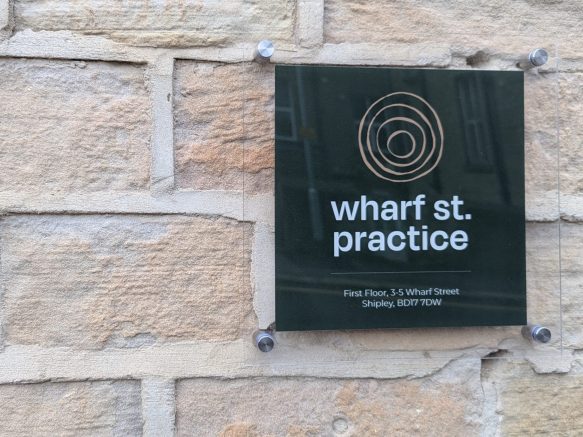 An image of the sign outside Wharf St. Practice, featuring the organisation's logo on a stone wall.